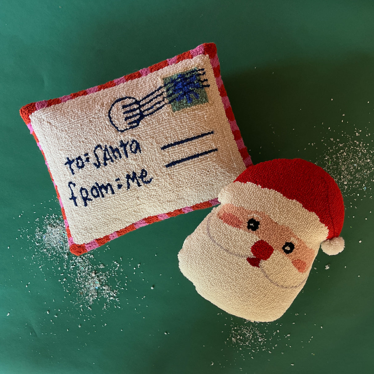 A hooked wool santa pillow and a pillow in the shape of a letter addressed to santa on a green background. 