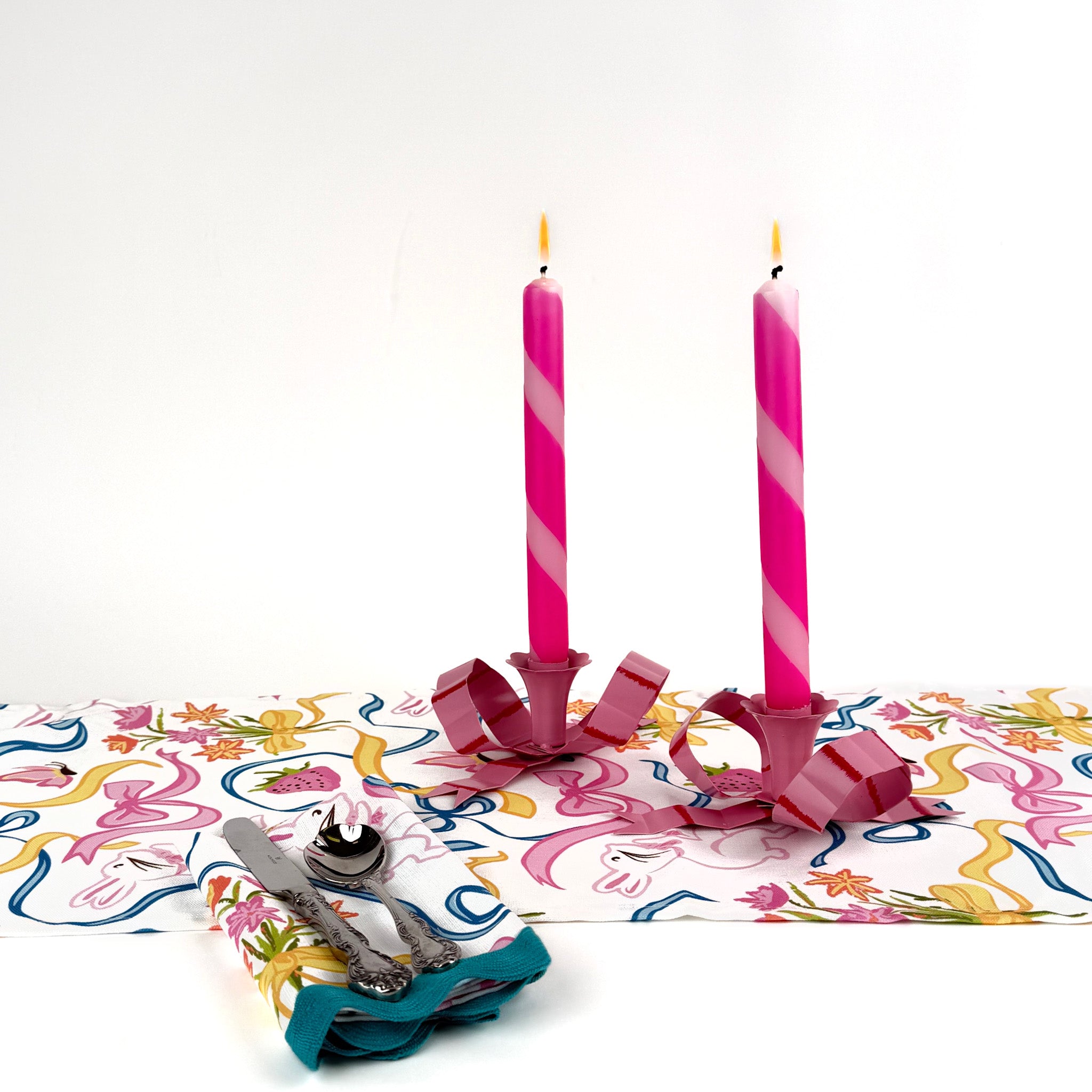 a pair of pink bow candle holders with lighted pink and white stripe candles on top of a bunny themed table runner and a dinner napkin cloth
