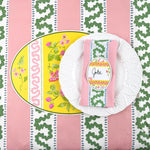 Table setting with a white plate, napkins, and name tags on a pink and green patterned tablecloth with an easter-egg-shaped-placemat