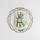 A white tole metal christmas charger with a pink ribbon and holly motifs around the edges against a white background and a napkin and plate placed on top of it