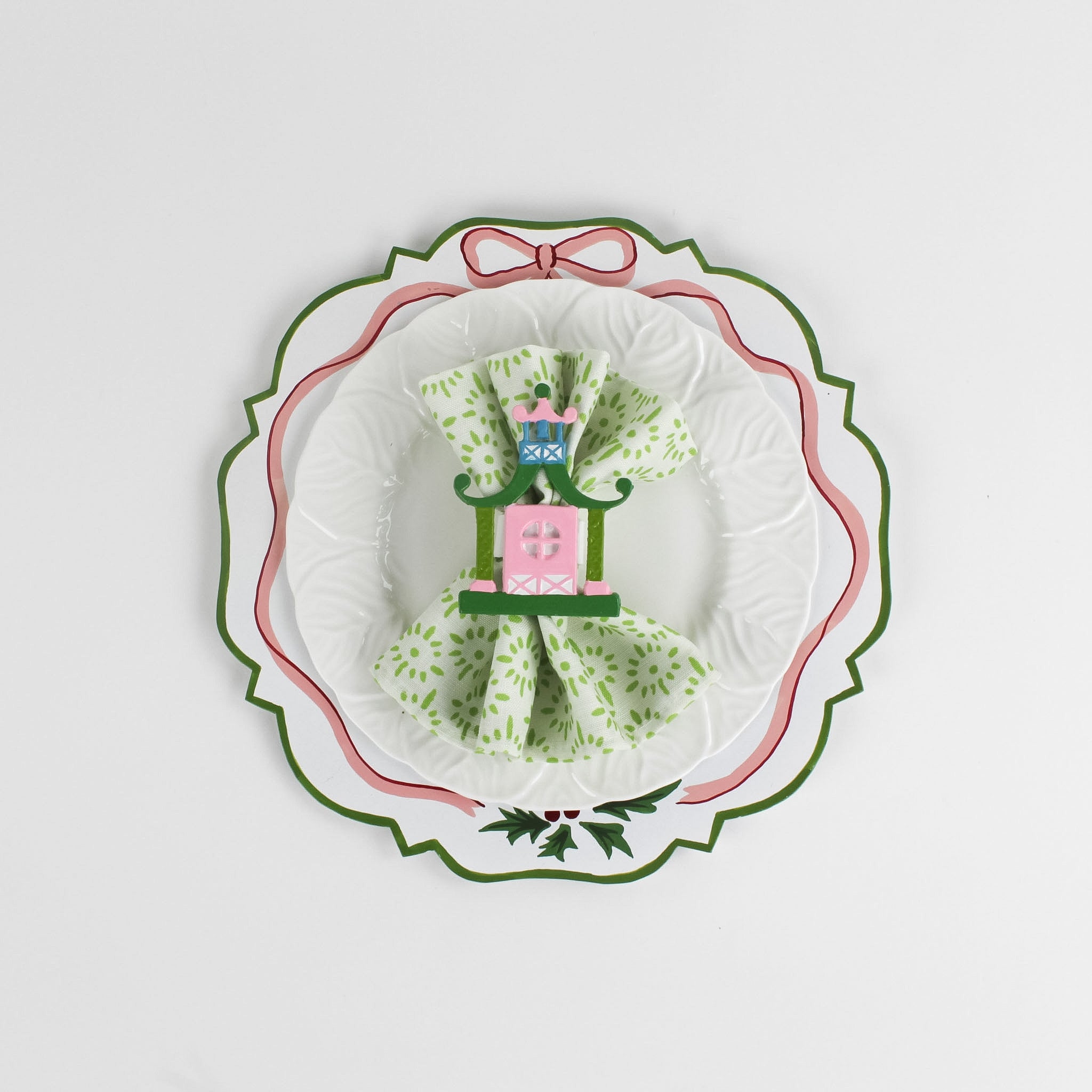 A white tole metal christmas charger with a pink ribbon and holly motifs around the edges against a white background and a napkin and plate placed on top of it