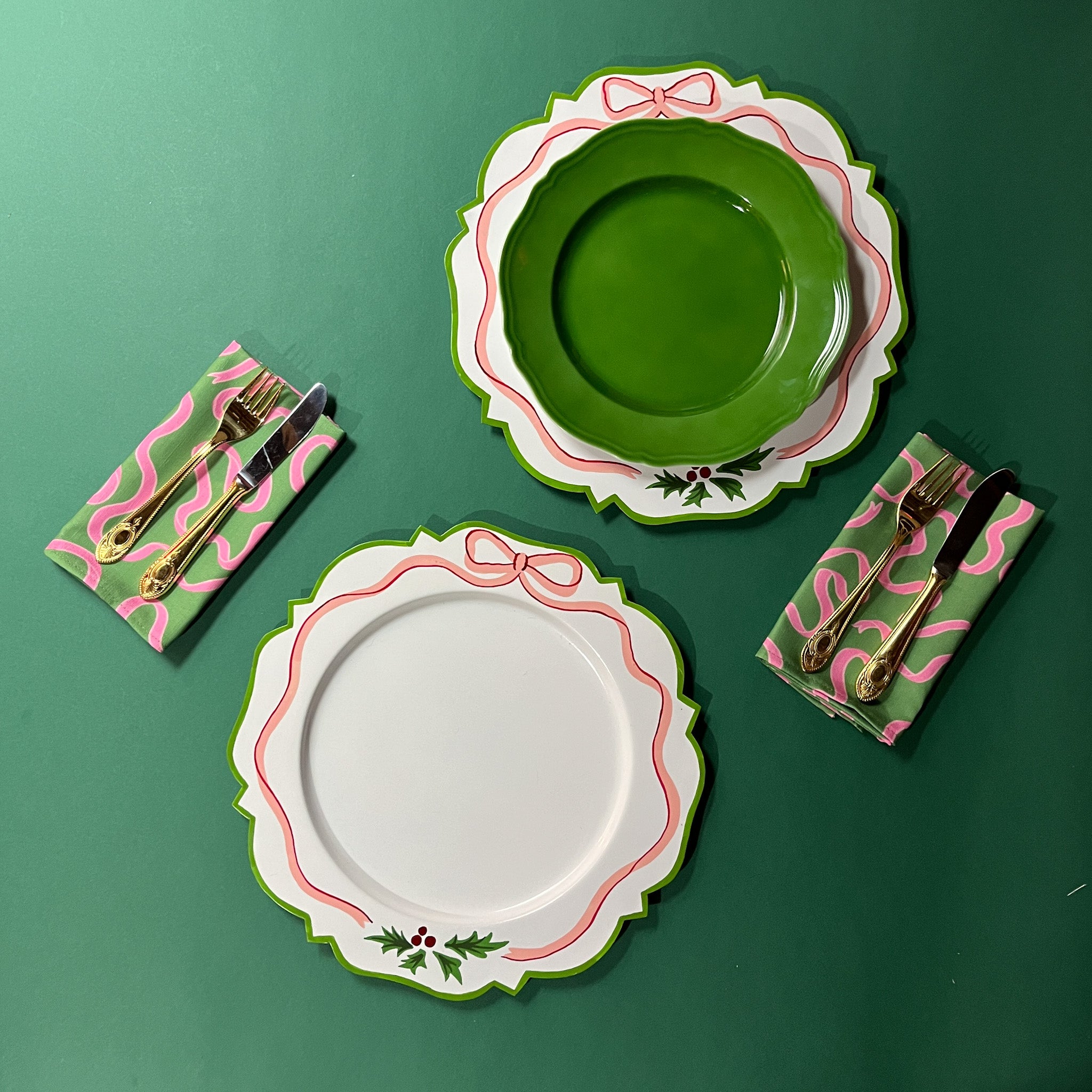 a pair of A white tole metal christmas charger plates with a pink ribbon and holly motifs around the edges against a green background with napkins and cutlery around it. 