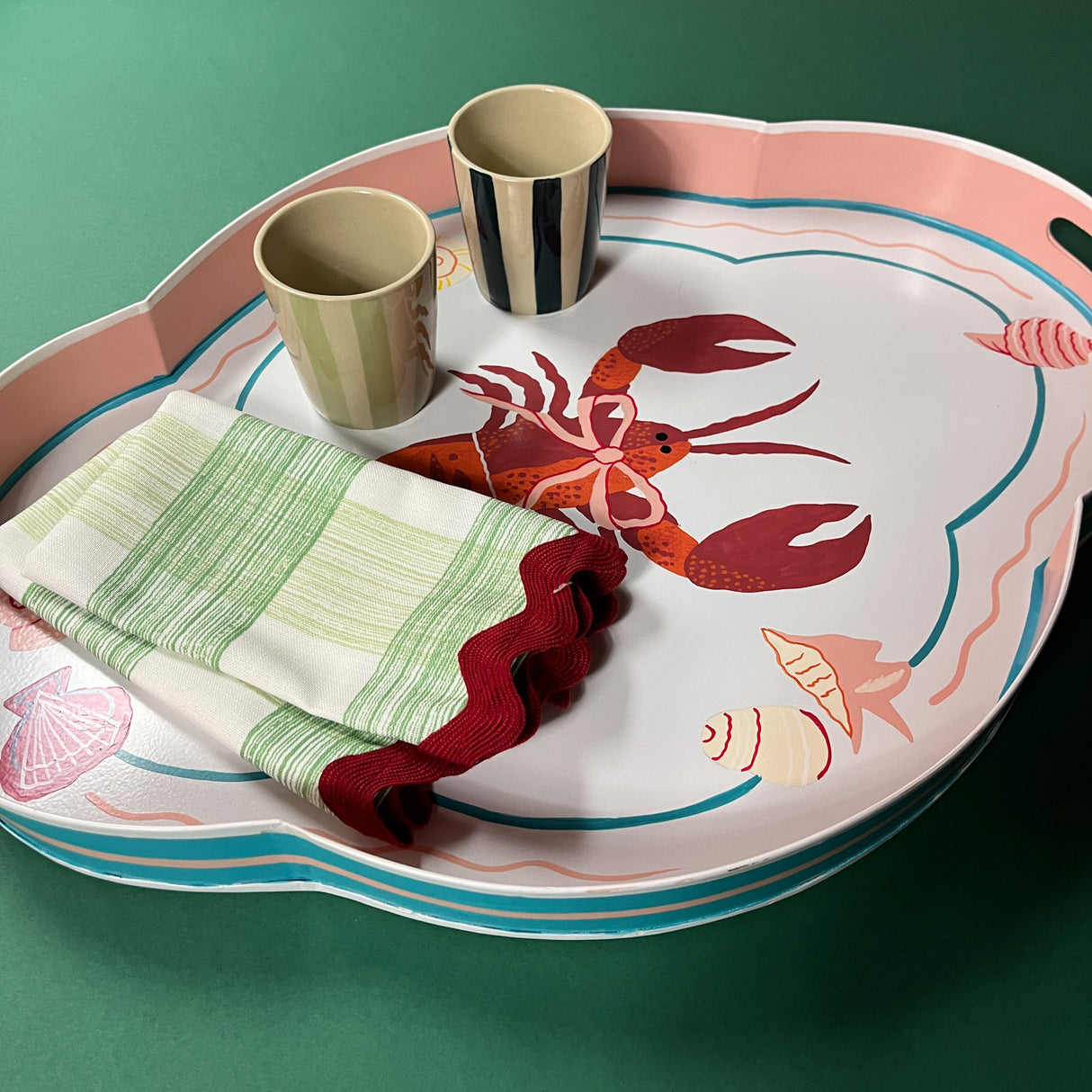 Lifestyle image of rock lobster pink and green tole metal tray with two ceramic cups and two Gin Lane dinner napkins with ric rac