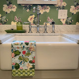 a floral tea towel over a white sink with a backround of green floral wallpaper.