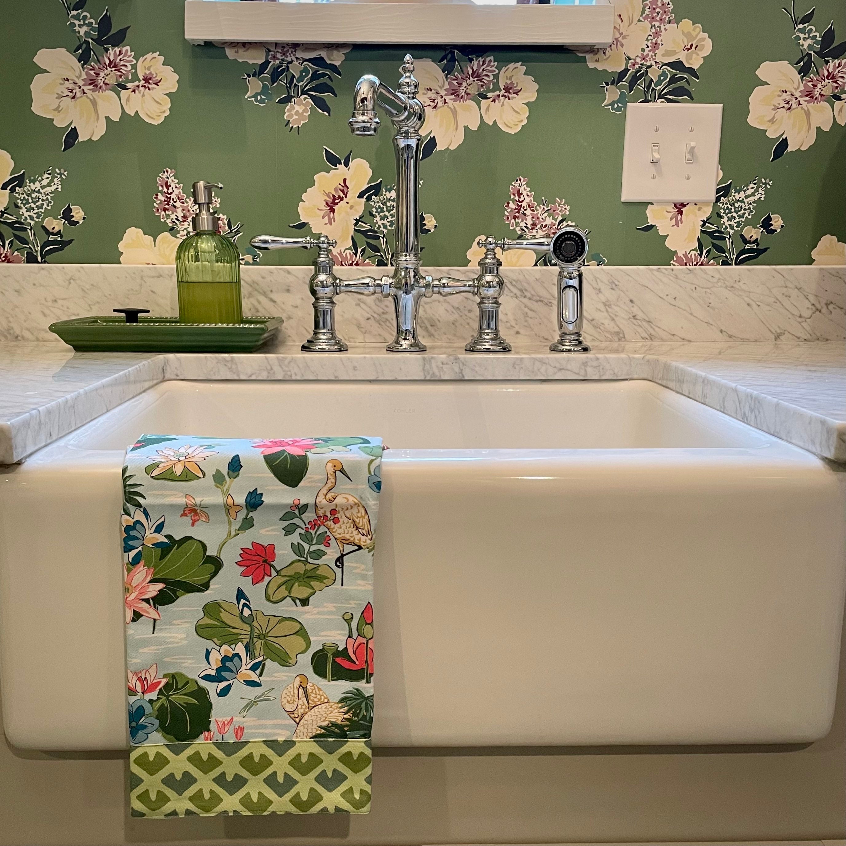 a floral tea towel over a white sink with a backround of green floral wallpaper.