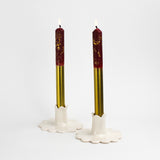 Two lit gold and red taper candles displayed in white scalloped ceramic candle holders.