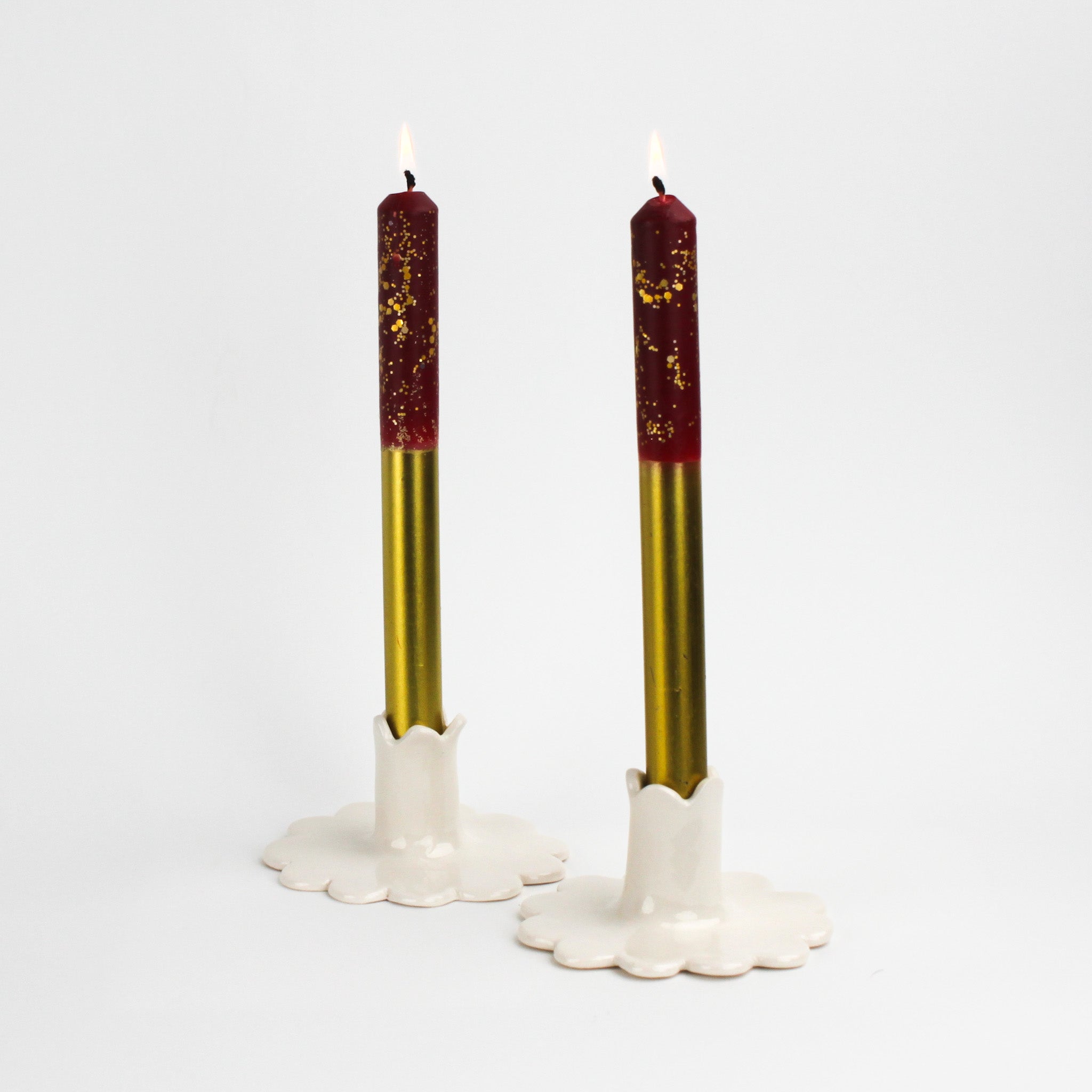 Two lit gold and red taper candles displayed in white scalloped ceramic candle holders.