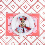 A styled coastal table setting featuring a lobster themed paper placemat, white plate, nautical napkin with anchor ring, and coral trellis patterned background.