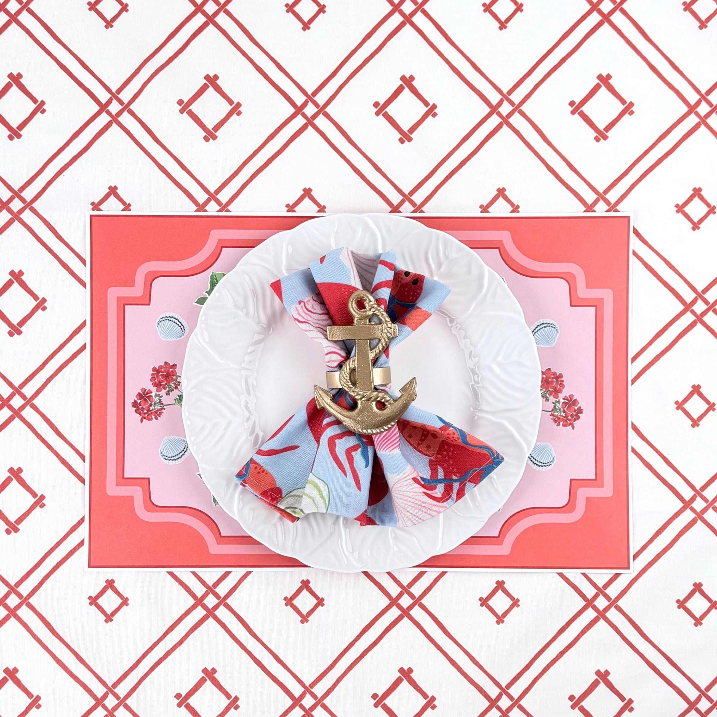 A styled coastal table setting featuring a lobster themed paper placemat, white plate, nautical napkin with anchor ring, and coral trellis patterned background.