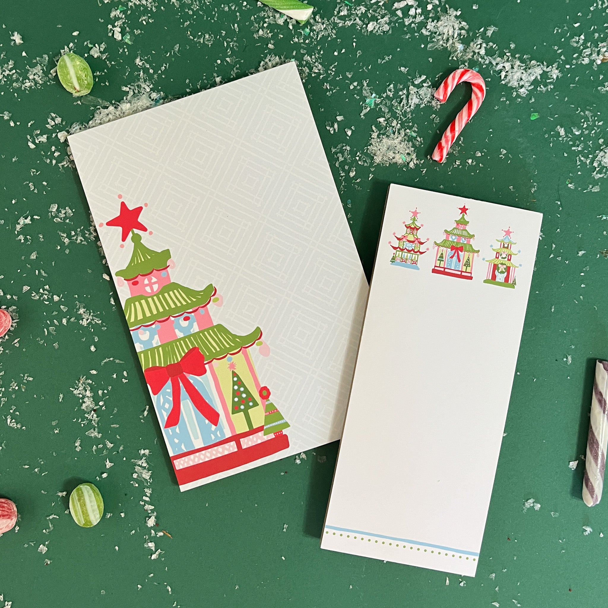 Two christmas notepads with festive pagoda illustrations, including trees and presents, placed on a green surface with candy canes and faux snow. 
