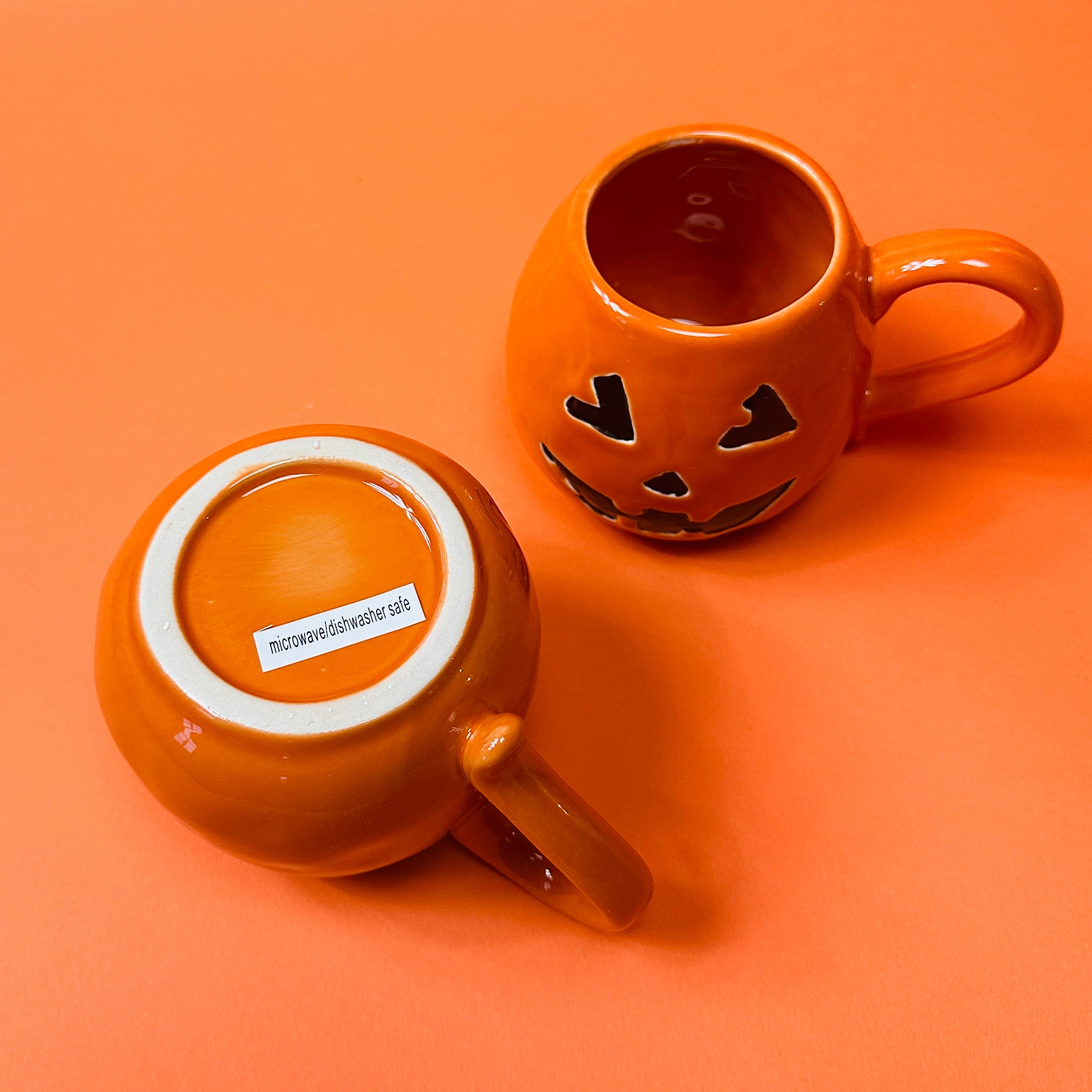 Orange ceramic mugs with a jack o'lantern design, one with a handle, one showing the bottom and the other is the top.
