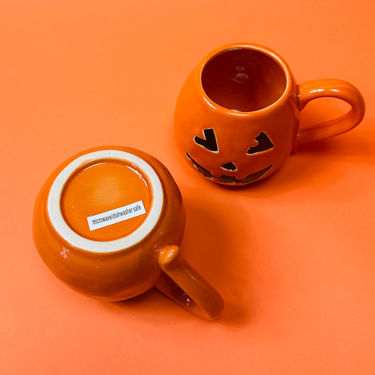 Orange ceramic mugs with a jack o'lantern design, one with a handle, one showing the bottom and the other is the top.