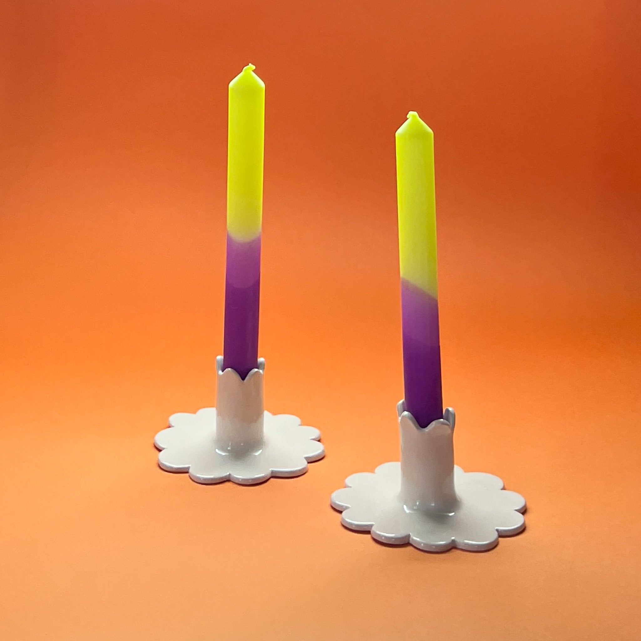 Two taper candles with a purple and green gradient, standing in white candle holders against an orange background.