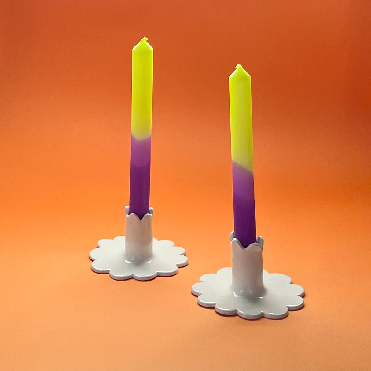 Two taper candles with a purple and green gradient, standing in white candle holders against an orange background.