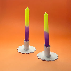 Two taper candles with a purple and green gradient, standing in white candle holders against an orange background.
