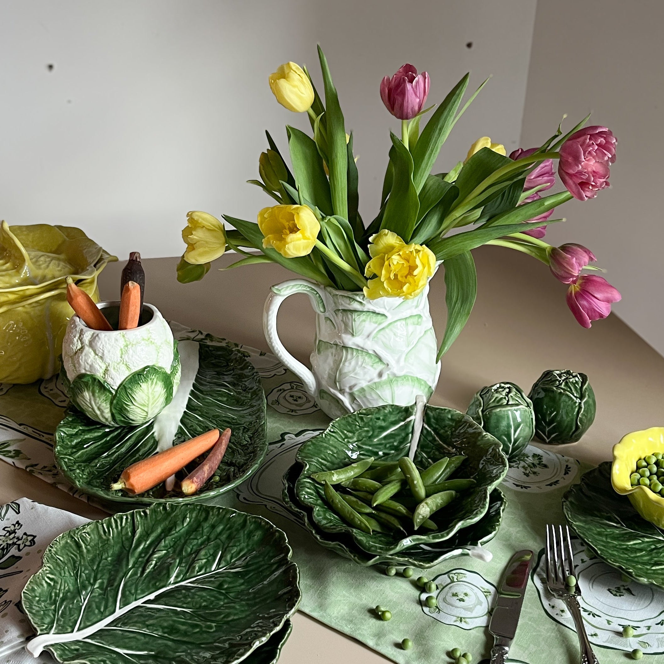 Spring tabletop arrangement with cabbage-leaf ceramic dishes, fresh vegetables, and yellow tulips in a green-toned setting.