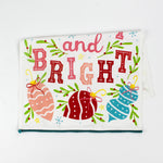 colorful christmas tea towel with with ornament motifs, blue pom pom trim and text that reads "Merry and Bright" against a white background. 