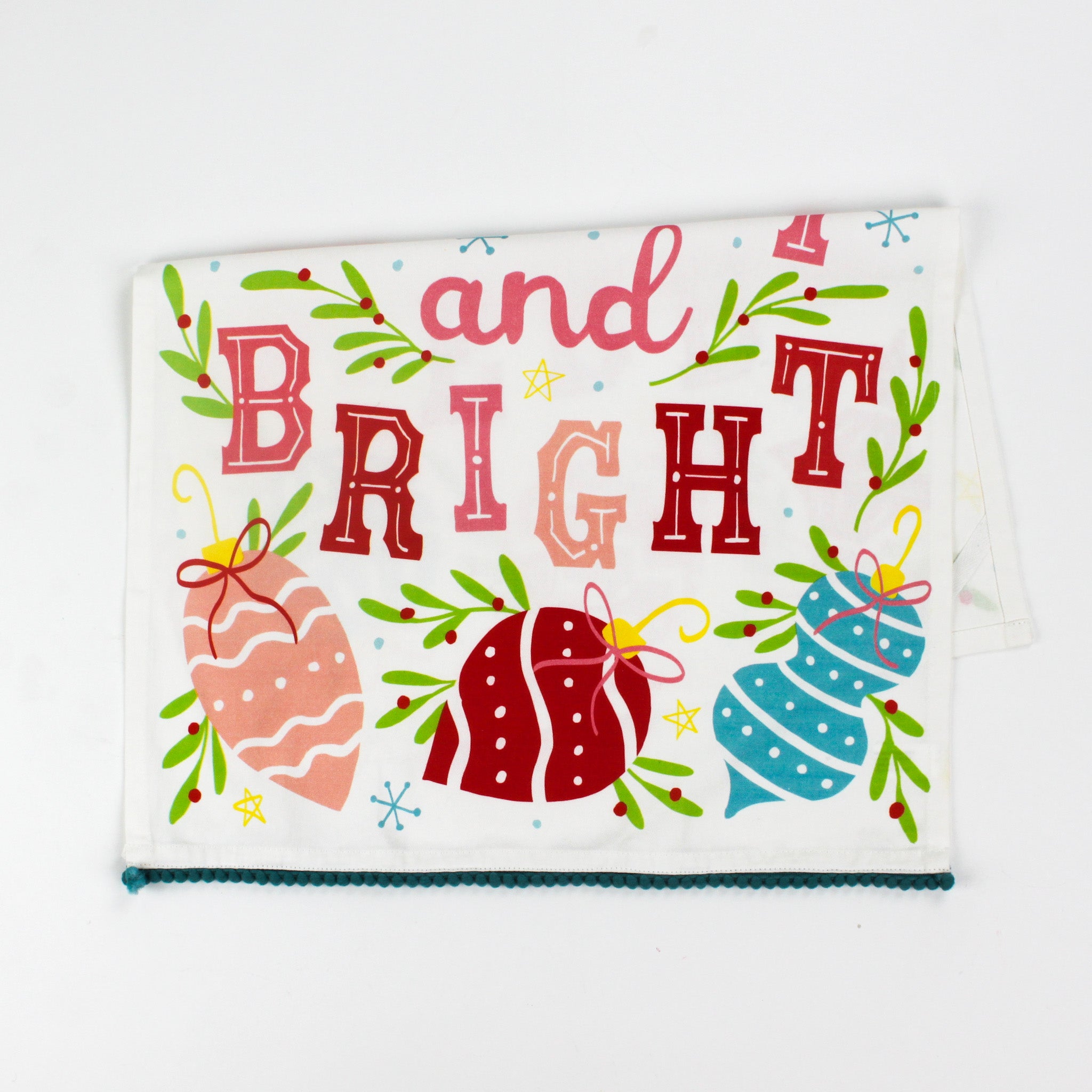 colorful christmas tea towel with with ornament motifs, blue pom pom trim and text that reads "Merry and Bright" against a white background. 