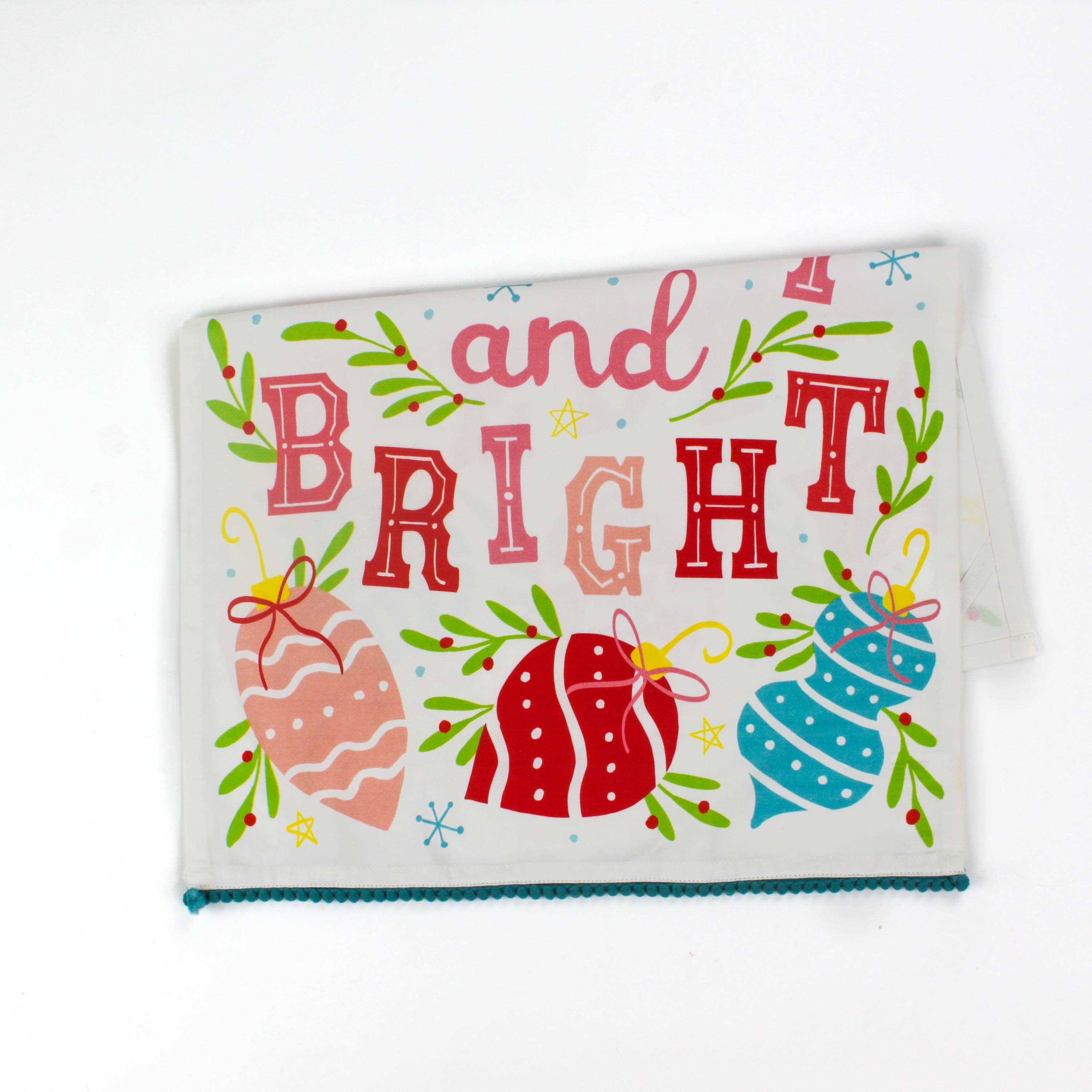 colorful christmas tea towel with with ornament motifs, blue pom pom trim and text that reads "Merry and Bright" against a white background. 