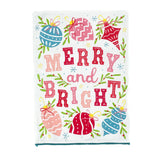 colorful christmas tea towel with with ornament motifs, blue pom pom trim and text that reads "Merry and Bright" against a white background. 