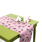 Pink holiday-themed table runner on a green table styled with two festive mugs filled with marshmallows and candy canes.
