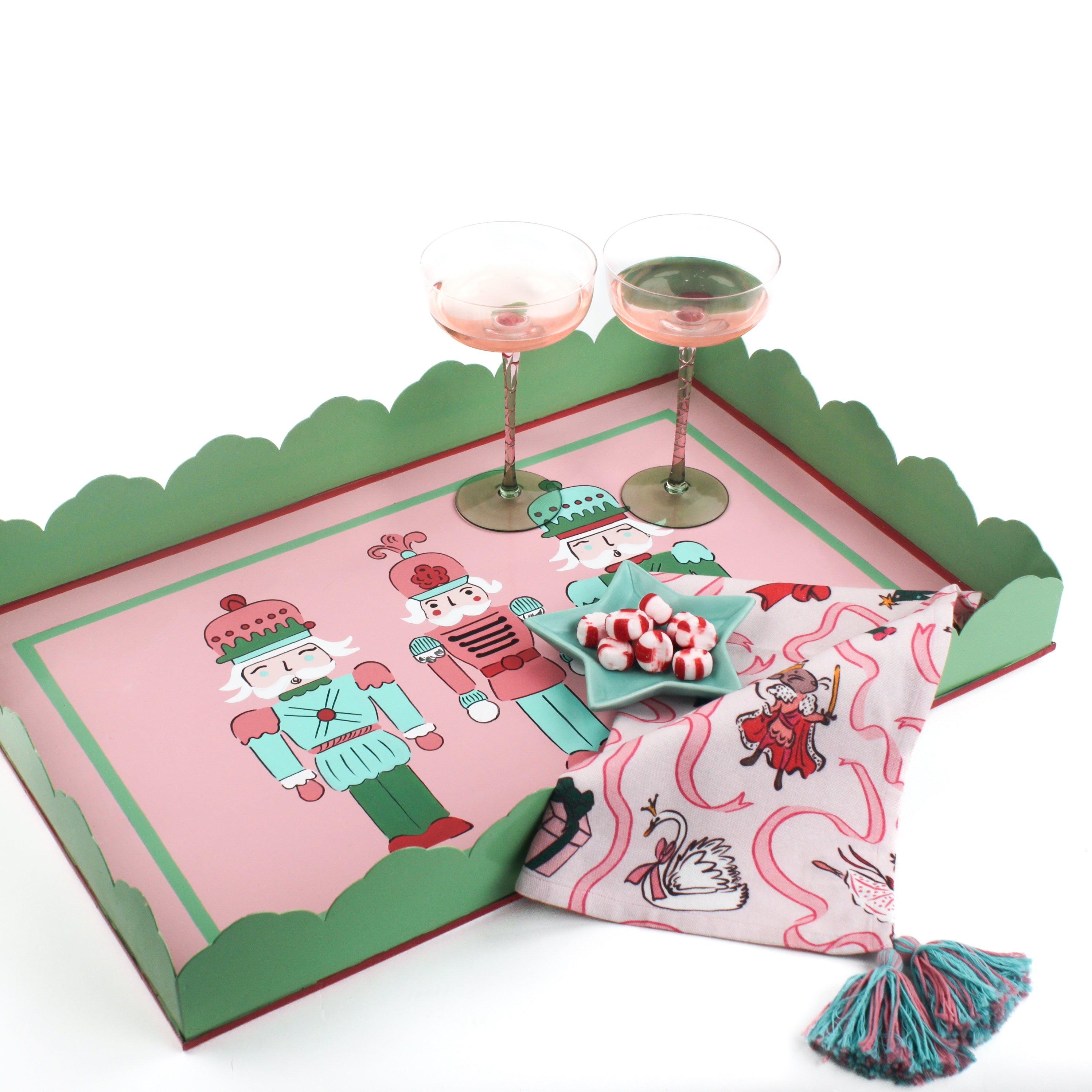 A pink and green nutcracker christmas tray filled with a pink christmas napkin, a blue star dish and two pink champagne coupes. 