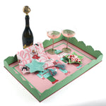 A pink and green nutcracker christmas tray filled with a pink christmas napkin, a blue star dish, two pink champagne coupes and a bottle of champagne.