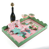 A pink and green nutcracker christmas tray filled with a pink christmas napkin, a blue star dish, two pink champagne coupes and a bottle of champagne.
