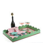 A pink and green nutcracker christmas tray filled with a pink christmas napkin, a blue star dish, two pink champagne coupes and a bottle of champagne.