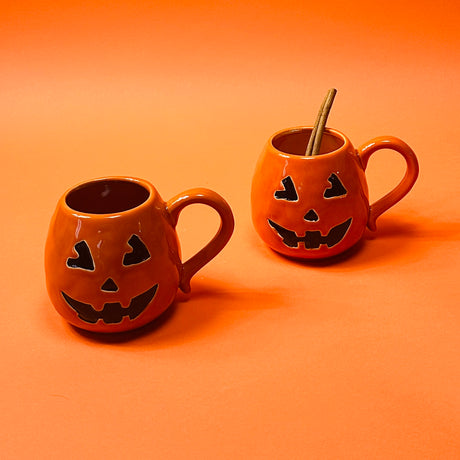 Two ceramic mugs with a jack o'lantern design, one with a handle, on an orange background.
