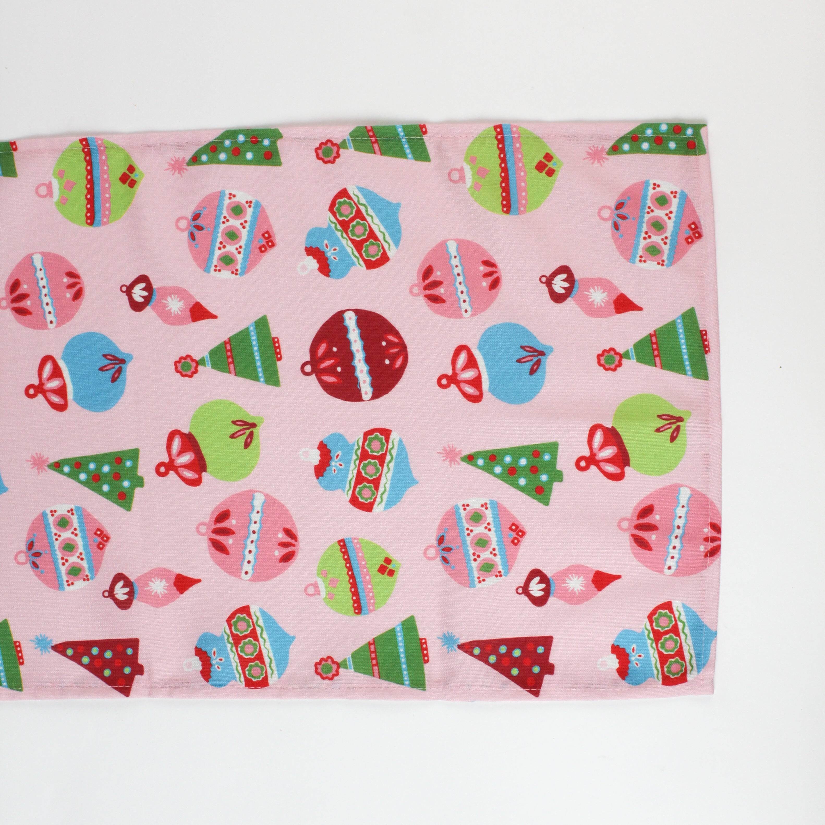 a pink christmas table runner featuring a pattern of colorful ornaments on a white background. 