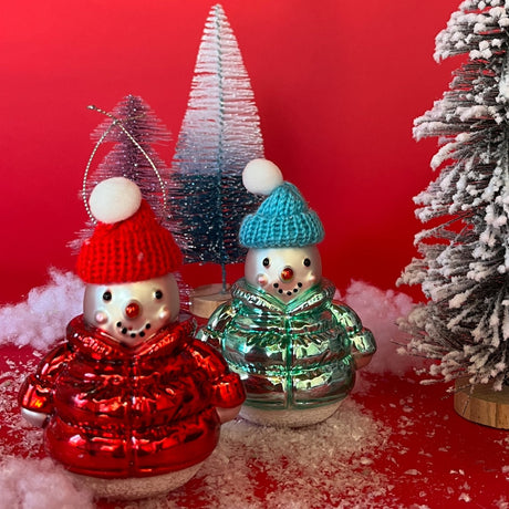 Two glass Christmas snowman ornaments with knitted caps, one red and one green, standing on a faux snow surface with artificial tree branches around. The red snowman is topped with a pom-pom, and both are surrounded by falling snowfake.