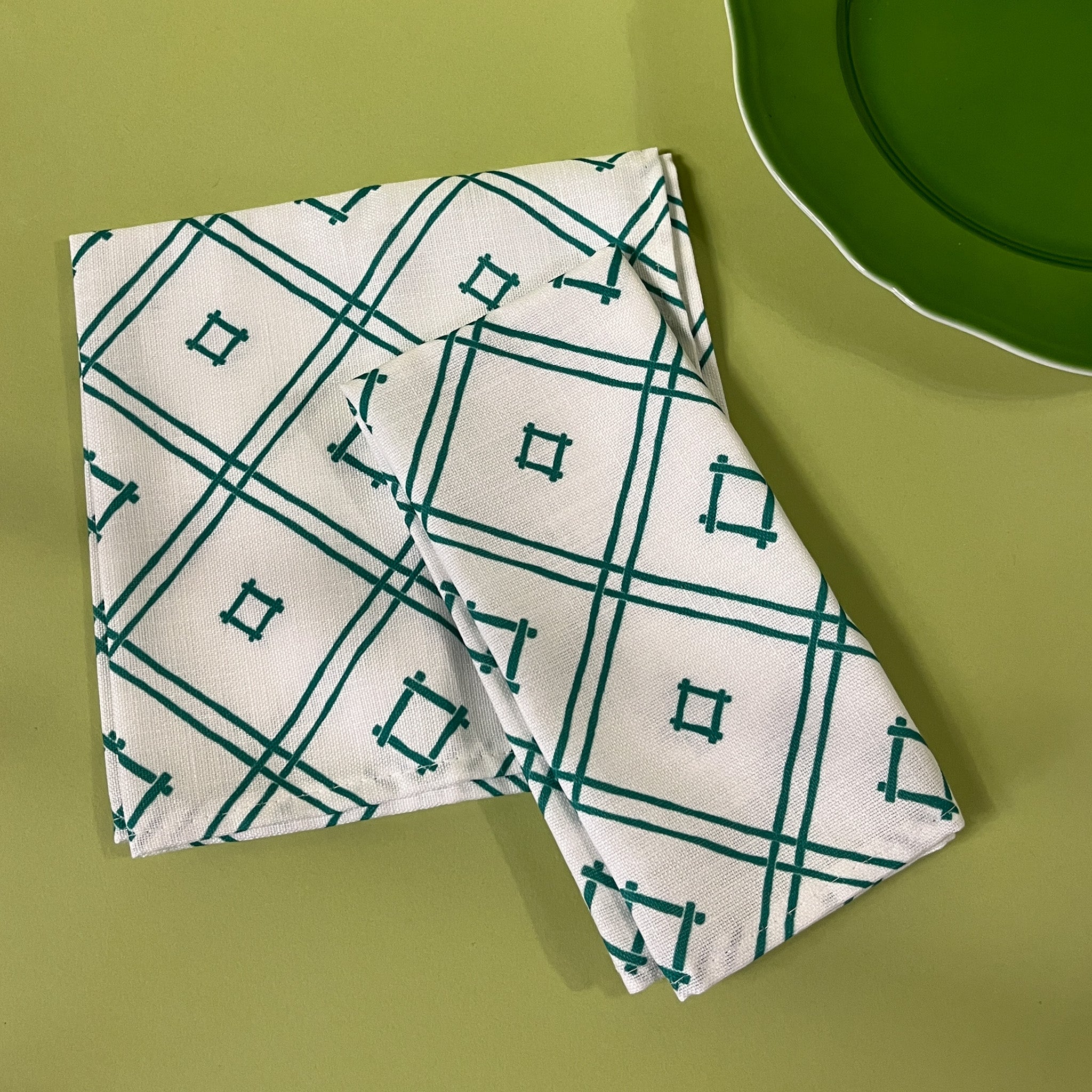 a pair of white and green cloth dinner napkins in Island House Small trellis print madcap cottage fabric against a light green background next to a green plate

