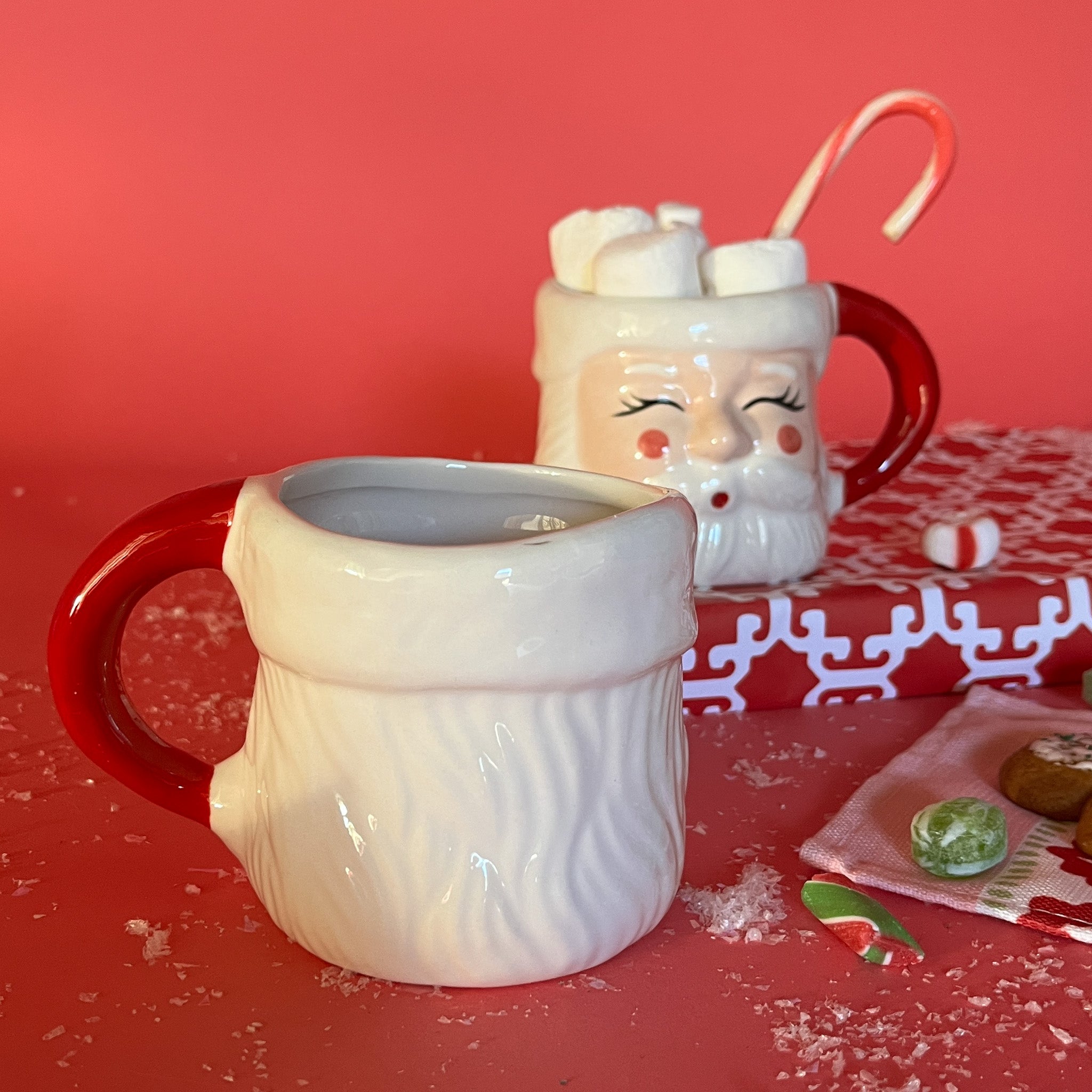 reverse side of a ceramic Christmas santa mug with a jolly santa face design with red cheeks and a red handle on a red, christmas themed background with christmas candy and wrapped gifts. 