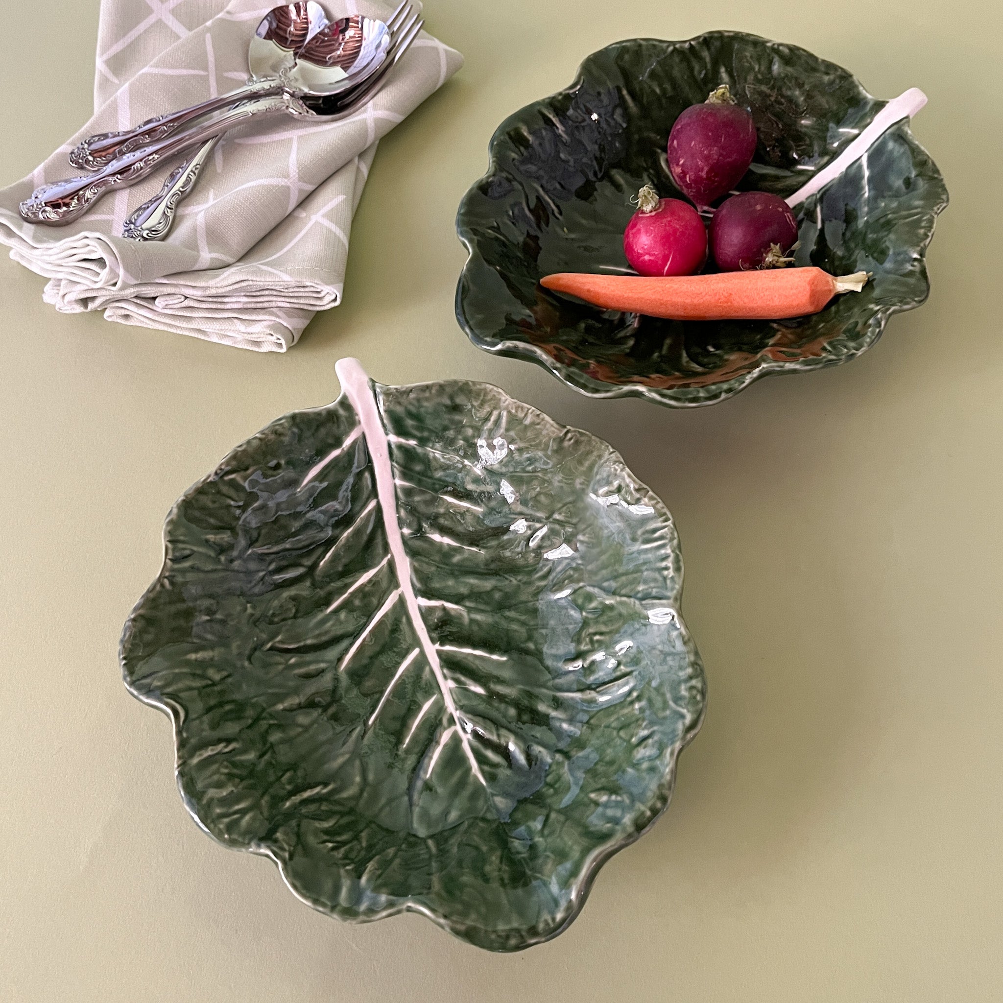 two ceramic cabbage leaf bowls, next to napkins and cutlery with one bowl filled with radishes 