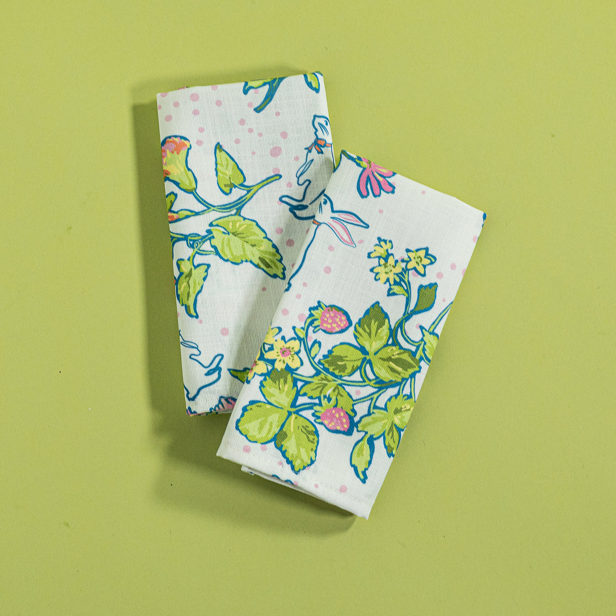 A pair of multicolor bunny and strawberry print fabric Easter napkins against a green background. 
