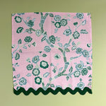 An open, flat pink and green chinoiserie cloth dinner napkin in temple garden print  madcap cottage fabric with dark green ric rac trim against a light green background