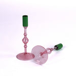 Pair of pink glass taper candle holders with turned stems and round bases, each fitted with a green candle sleeve, photographed on a white background.