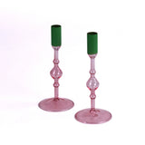 Pair of pink glass taper candle holders with turned stems and round bases, each fitted with a green candle sleeve, photographed on a white background.