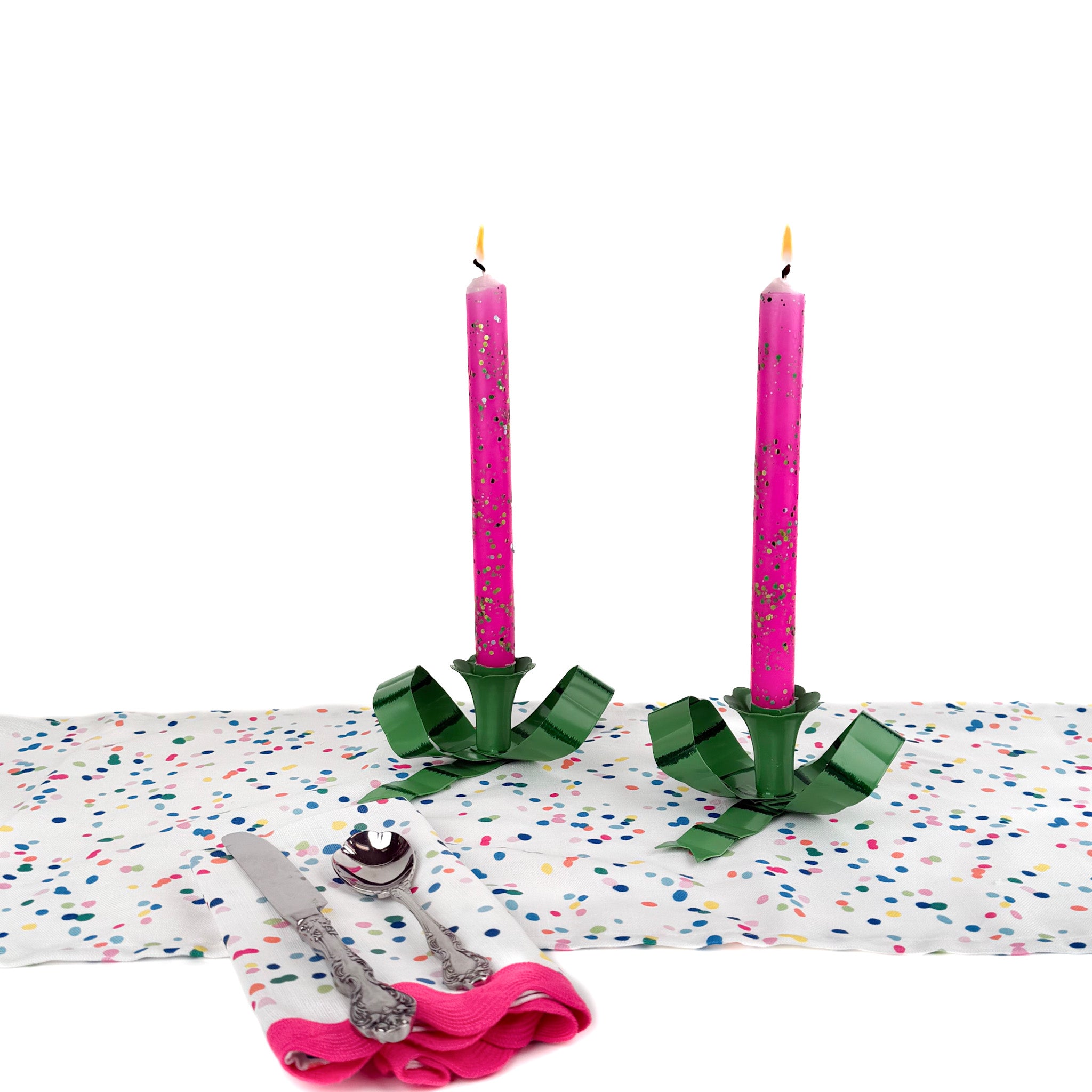 Pink glitter taper candles lit on a colorful confetti-pattern table setting with green candle holders and silverware.