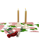 Green and pink pinstripe taper candles lit on a spring tablespace with strawberry-pattern table linens, silverware, and decorative candle holders.