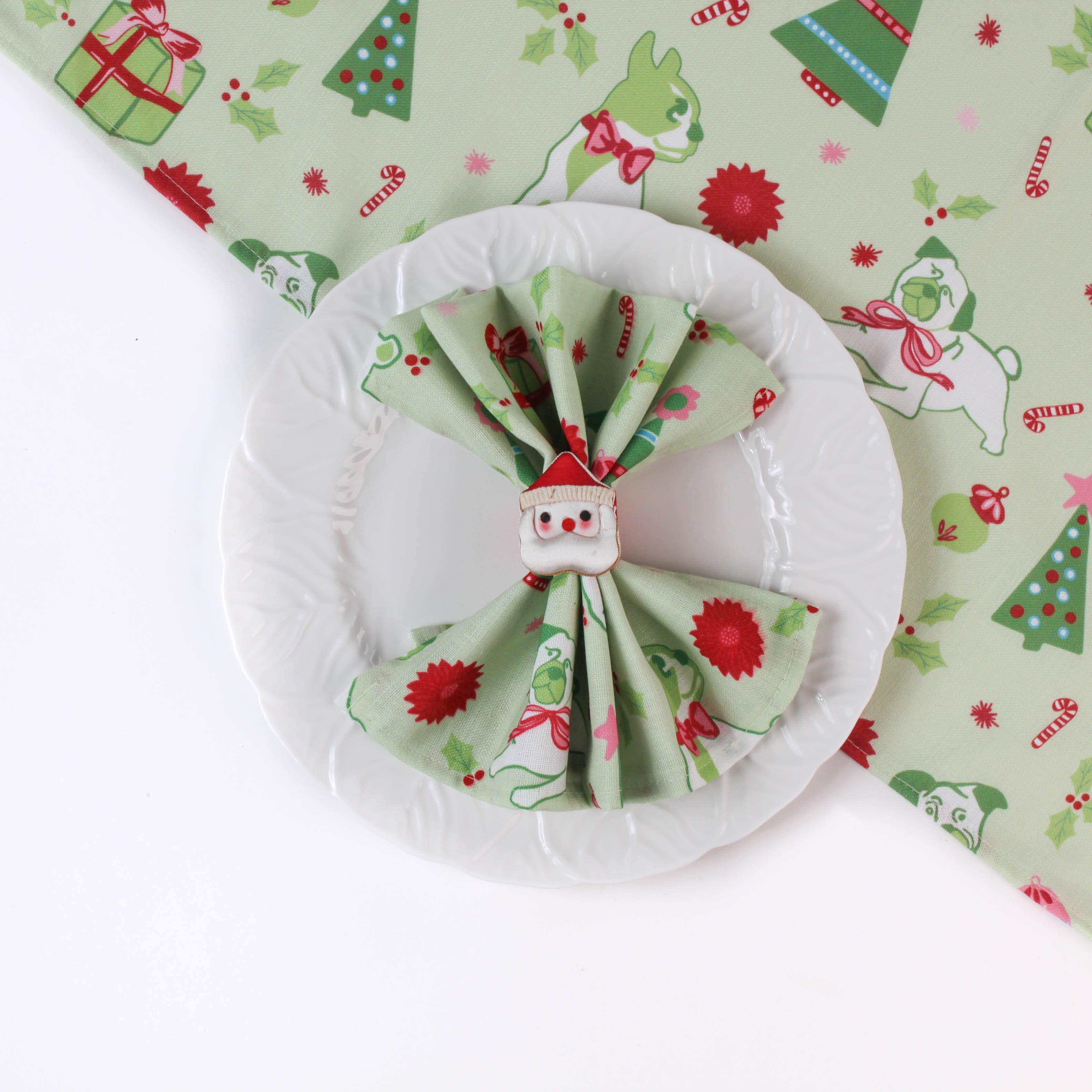 a green christmas napkins with pugs, christmas trees and candy cane motifs inside a santa napkin on top of a matching table runner on a white background