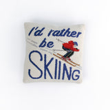 Square decorative pillow with a skier in a red jacket and blue helmet on snowy slopes, featuring the phrase “I’d rather be SKIING” in bold blue text.
