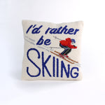 Square decorative pillow with a skier in a red jacket and blue helmet on snowy slopes, featuring the phrase “I’d rather be SKIING” in bold blue text.