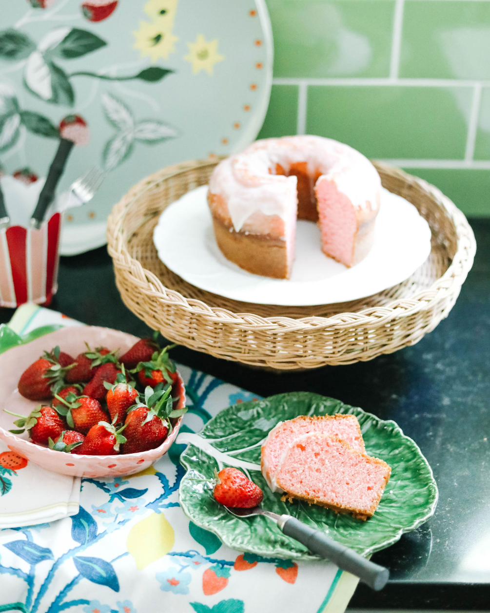 a rattan pedestal bowl holding a cake and surround by strawberry decor in a green kitchen. 