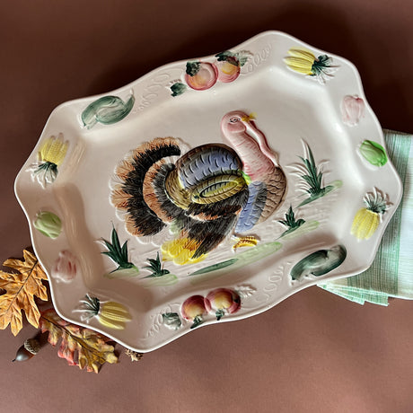 A vintage turkey platter with scalloped edges, featuring a three-dimensional turkey in the center surrounded by colorful vegetables and floral patterns on a brown background. The platter is displayed on a textured surface with a green and white napkin to the side.
