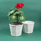 Two white ceramic planters with a wicker-style texture, one holding a faux red flowering plant, displayed on a green background