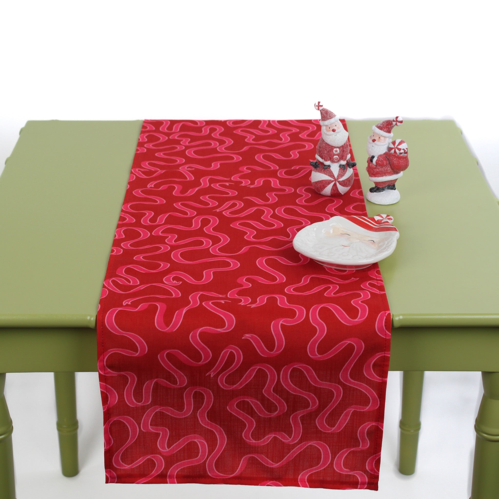 Red squiggle-pattern table runner styled on a light green table with two Santa figurines and a white dish.