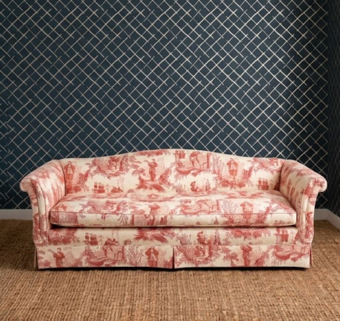 A classic three-seat sofa upholstered in a red and cream toile de Jouy pattern, set against a dark navy blue diamond-patterned wallpaper and a woven jute rug.