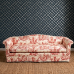 A classic three-seat sofa upholstered in a red and cream toile de Jouy pattern, set against a dark navy blue diamond-patterned wallpaper and a woven jute rug.